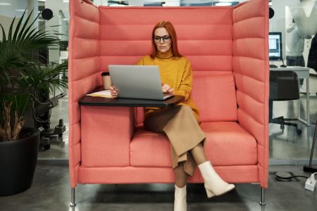Businesswoman in a yellow sweater working on a laptop in a modern office booth setting, exhibiting style and focus.